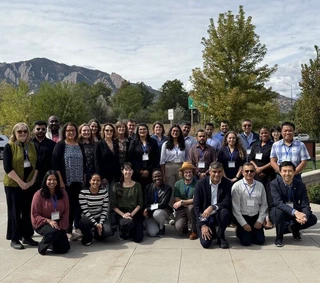About thirty meeting participants gathered outdoors for a group photograph on a sunlit plaza, standing and kneeling in two rows and wearing conference lanyards, with autumn trees and the Flatirons rising behind them.