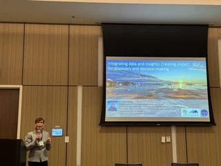 Twila Moon speaking with a handheld microphone beside a projection screen showing her title slide, which reads Integrating data and insights: Creating impact for discovery and decision making over a photograph of icebergs on a calm sea at low sun.