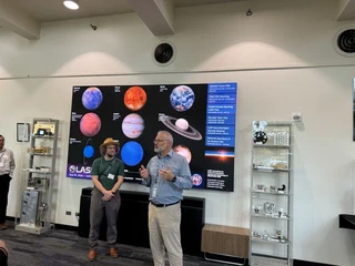 Two LASP staff members speaking to visitors in an exhibition space, standing in front of a large display of planetary images and beside cases of spaceflight instrument hardware. A schedule board on the display lists the iHARP meeting LASP tour.